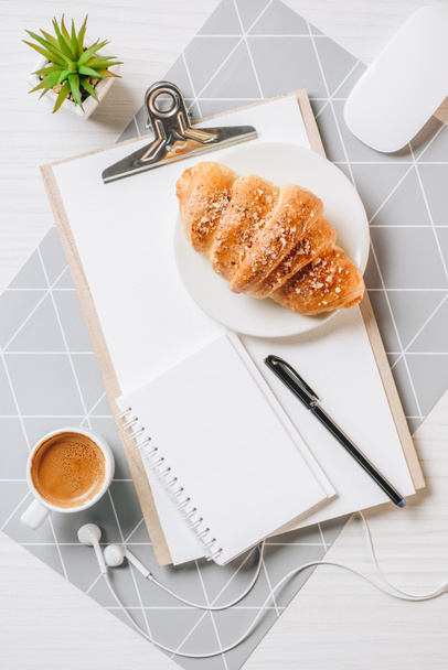 view from above of empty clipboard, earphones, croissant and coffee cup at table in office  - Zdjęcie, obraz