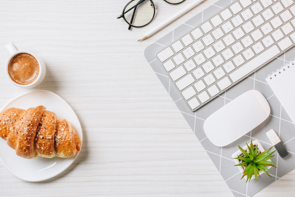 top view of arranged computer keyboard, eyeglasses, coffee cup and croissant at table in office  - Photo, Image