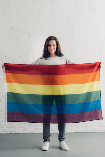 счастливая трансгендерная женщина с флагом гордости перед стеной из белого кирпича - Фото, изображение