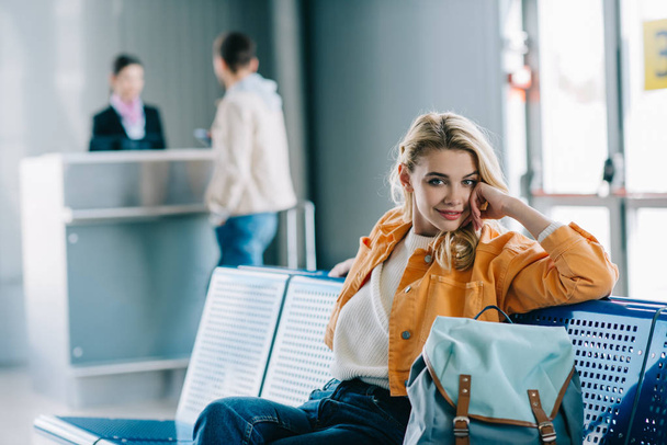 空港ターミナルに座ってカメラに笑顔のバックパックと魅力的な若い女性  - 写真・画像