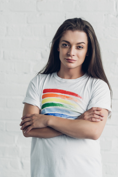 young transgender woman in white t-shirt with pride flag looking at camera with crossed arms in front of white brick wall - Photo, Image