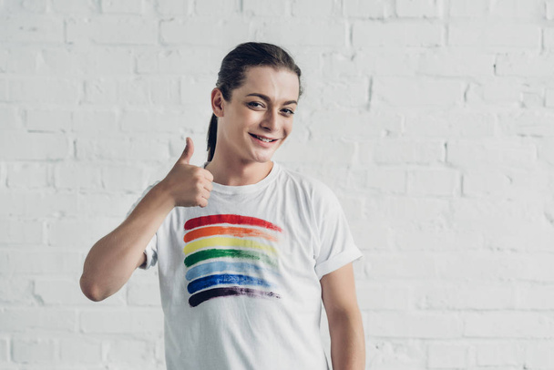 白いレンガの壁の前に親指を示す誇りフラグと白いTシャツの幸せなトランスジェンダーの女性 - 写真・画像
