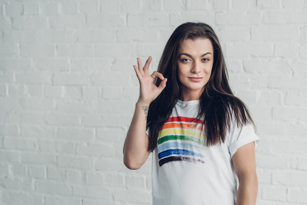 young transgender woman in white t-shirt with pride flag showing okay sign in front of white brick wall - Фото, зображення