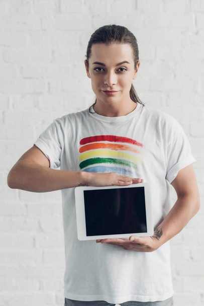 joven mujer transgénero en camiseta blanca con bandera de orgullo sosteniendo la tableta con pantalla en blanco delante de la pared de ladrillo blanco - Foto, Imagen