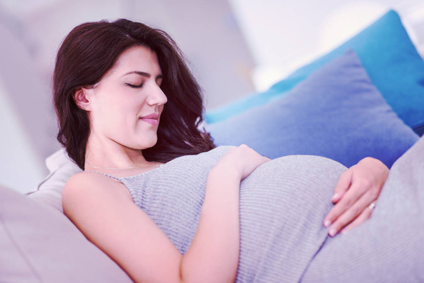 young happy pregnant woman keeping hand on belly while sitting on sofa at home - Foto, Imagem