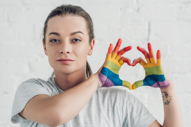 joven mujer transgénero haciendo el signo del corazón con las manos en colores de la bandera del orgullo delante de la pared de ladrillo blanco - Foto, imagen