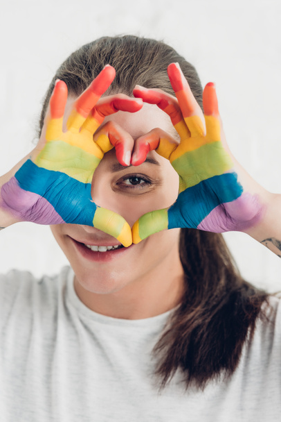 young transgender woman looking at camera while making heart sign with hands in colors of pride flag in front of white brick wall - Photo, Image