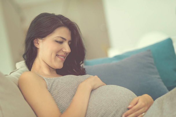 young happy pregnant woman keeping hand on belly while sitting on sofa at home - Foto, Imagen