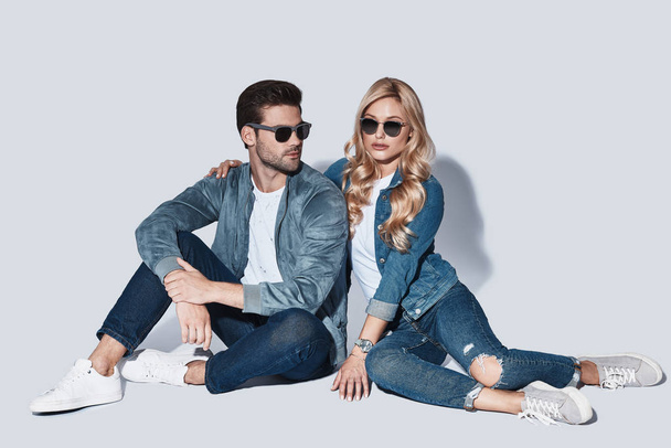 fashionable man and woman in denim wear sitting on floor in studio and wearing sun glasses, copy space - Фото, зображення