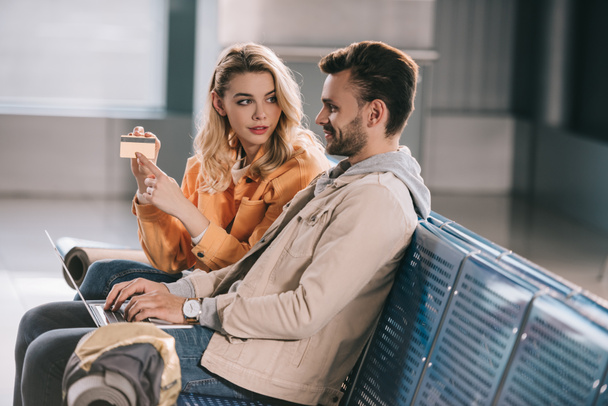 girl showing credit card to smiling man using laptop in airport - Foto, Imagen