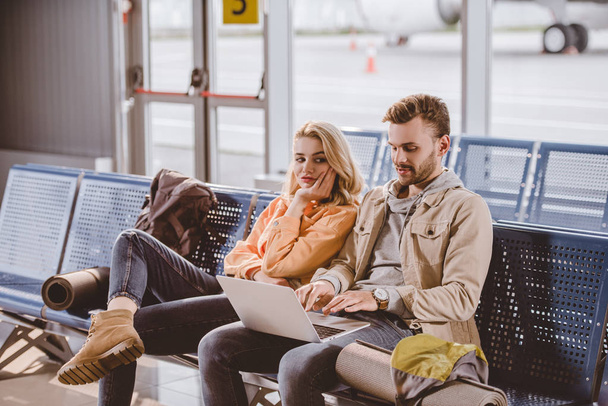 nuori pari matkustajat käyttävät kannettavaa tietokonetta ja odottavat lentoa lentokentän terminaalissa
 - Valokuva, kuva