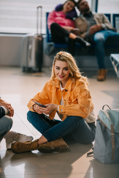 atractiva joven mujer sonriendo a la cámara mientras está sentada en el suelo y esperando el vuelo en el aeropuerto
 - Foto, Imagen