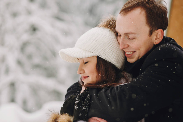 Κομψό ζευγάρι αγκαλιάζει στην ξύλινη καμπίνα στο φόντο το χιονισμένο δάσος στα βουνά. Ευτυχισμένη οικογένεια Αγκαλιάζοντας στο εξοχικό σπίτι στα βουνά του χειμώνα. Απόδραση διακοπές μαζί. Χώρο για το κείμενο - Φωτογραφία, εικόνα