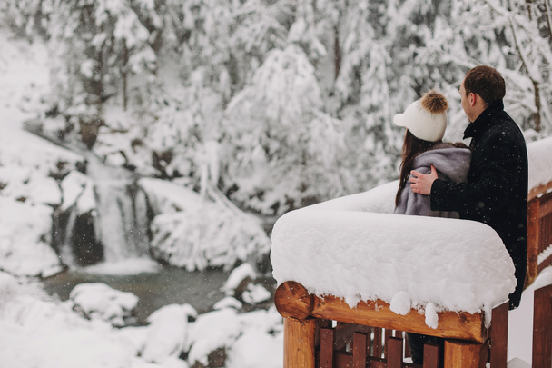 Stylish couple hugging and looking at snowy woods from wooden porch in mountains. Happy family relaxing on background of winter mountains. Holiday getaway together. Space for text - Photo, Image