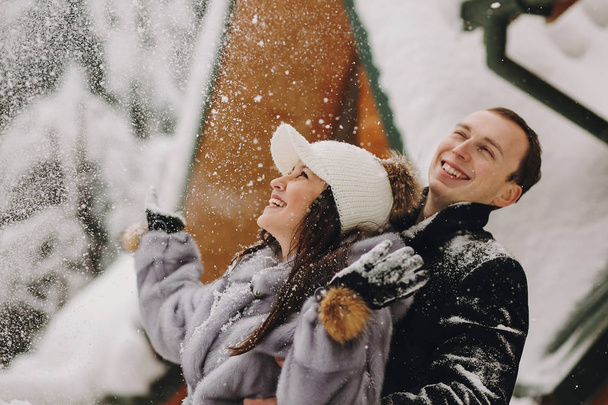 Stylish couple playing with snow in wooden cabin on background of winter snowy mountains. Happy joyful family having fun and smiling in snow. Emotional funny moments together - Photo, Image