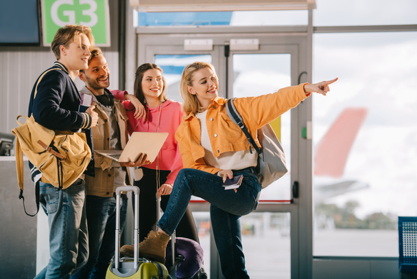 souriant jeunes amis à l'aide d'un ordinateur portable et pointant du doigt en attendant le vol à l'aéroport
  - Photo, image