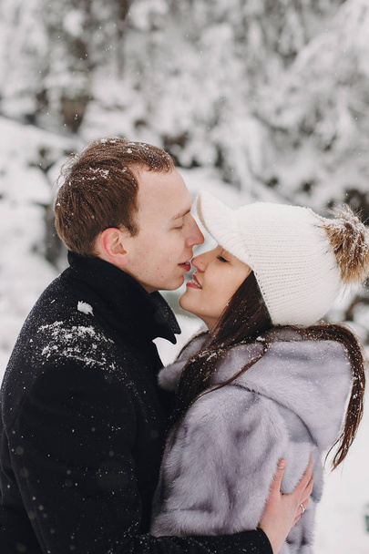 Stylish couple kissing in winter snowy mountains. Happy romantic man and woman in luxury clothes gently embracing at waterfall in snow. Holiday getaway together. Space for text - 写真・画像
