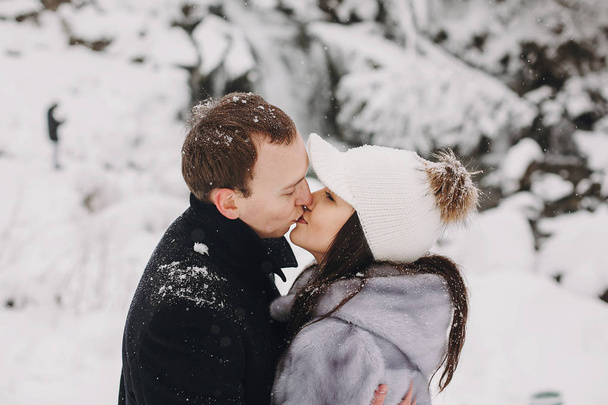 Stylish couple kissing in winter snowy mountains. Happy romantic man and woman in luxury clothes gently embracing at waterfall in snow. Holiday getaway together. Space for text - Foto, Imagen