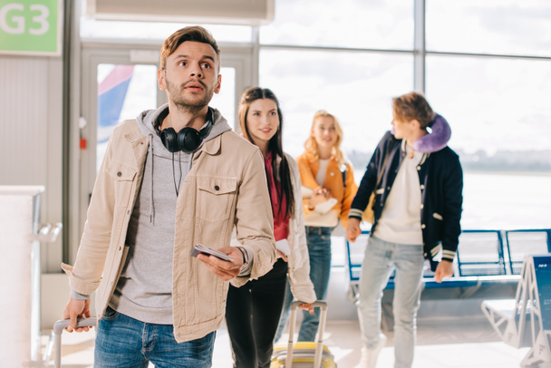 νεαρός άνδρας κρατά διαβατήριο και κοιτώντας ψηλά ενώ ταξιδεύετε με φίλους στο αεροδρόμιο  - Φωτογραφία, εικόνα