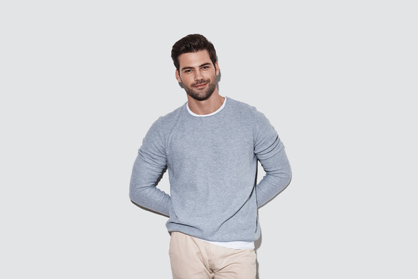 Charming man in casual wear looking at camera and smiling while standing against grey background - Foto, Bild