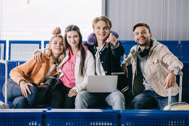 heureux jeunes amis utilisant un ordinateur portable et souriant à la caméra en attendant le vol dans le terminal de l'aéroport
   - Photo, image