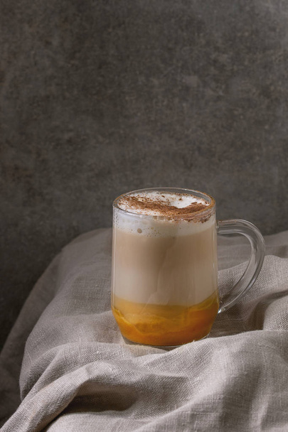 Glass of pumpkin layered spice latte with pumpkin puree, milk foam and cinnamon standing on crumpled table cloth with grey wall at background. Copy space - Fotoğraf, Görsel