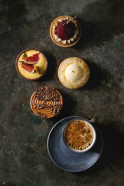 Variety of sweet tartlets with chocolate, caramel, pears, figs with cup of coffee on black texture background. Flat lay, space - Foto, Imagem