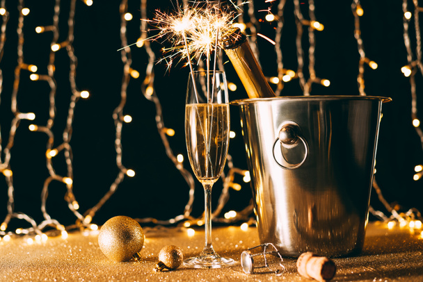 пляшка шампанського у відрі, склянка з різдвяними блискітками на світлому фоні гірлянди
 - Фото, зображення