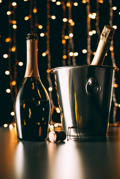 champagne bottle on table and bucket with bottle on garland light background, christmas concept - Photo, Image