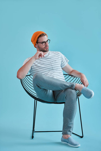 Handsome man isolated. Stylish young man in eyeglasses is  posing while sitting on chair on blue background. - Photo, image