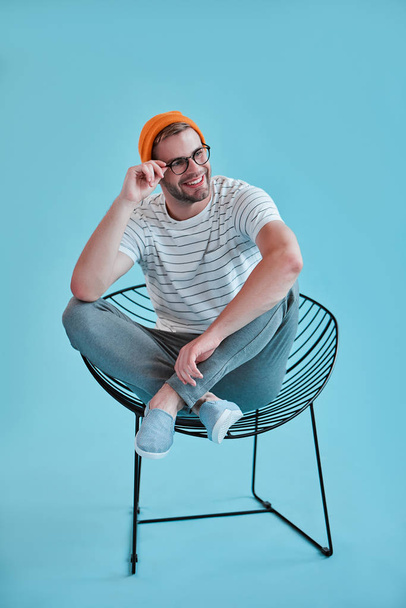 Handsome man isolated. Stylish young man in eyeglasses is  posing while sitting on chair on blue background. - Photo, Image