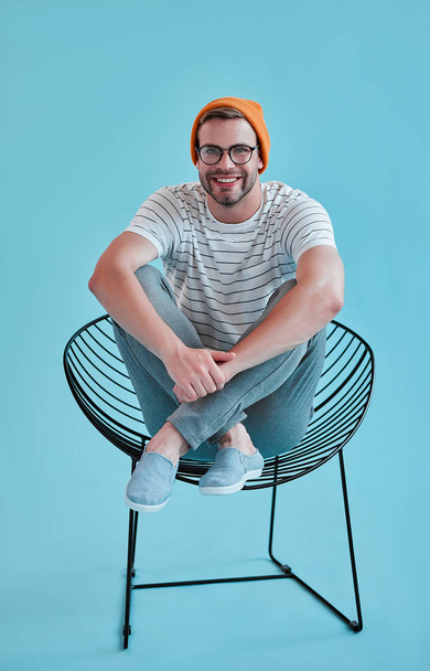 Handsome man isolated. Stylish young man in eyeglasses is  posing while sitting on chair on blue background. - Фото, изображение