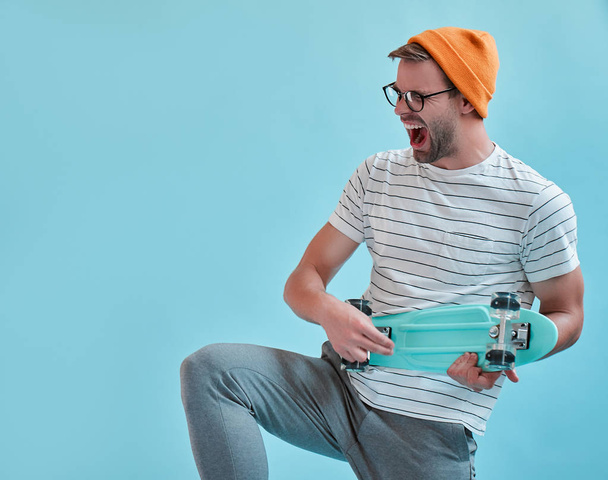 Um homem bonito isolado. Jovem elegante em óculos está posando com skate no fundo azul
. - Foto, Imagem