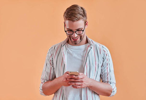 Handsome man isolated. Stylish young man in eyeglasses is  posing with smart phone on orange background. - Foto, Imagem