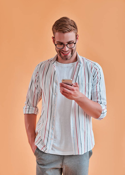Handsome man isolated. Stylish young man in eyeglasses is  posing with smart phone on orange background. - Foto, imagen