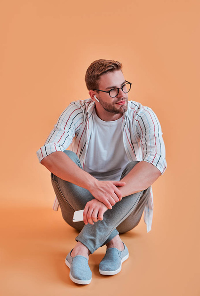 Handsome man isolated. Stylish young man in eyeglasses is  posing with smart phone on orange background. Listening to music using wireless earphones - Фото, зображення