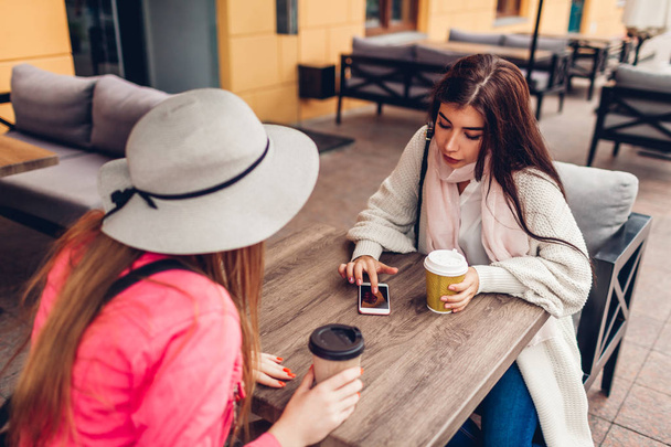 Two women chatting while having coffe in outdoor cafe. Happy friends using phone checking pictures. Girls hang out and having fun - Photo, Image