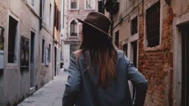Back view of confident stylish young businesswoman in hat and suit walking along narrow ancient street in Venice, Italy. - Video