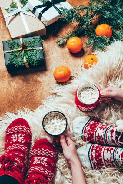 vista parcial de las mujeres en calcetines con tazas de bebidas de cacao en el fondo con mandarinas y regalos de Navidad envueltos
 - Foto, Imagen
