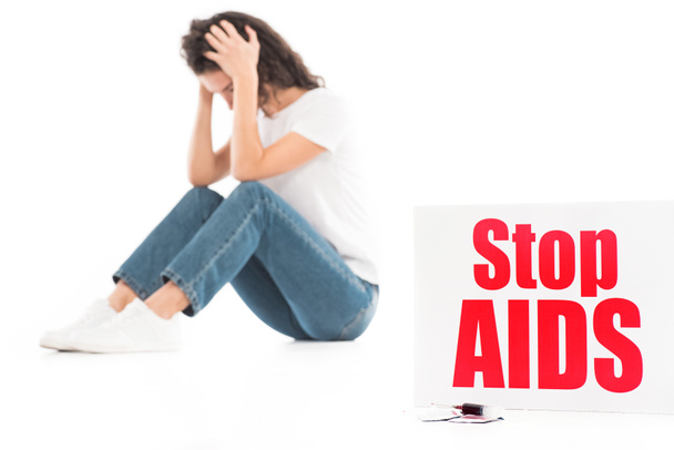 sad woman sitting and touching head isolated on white, stop aids card on foreground - Photo, Image