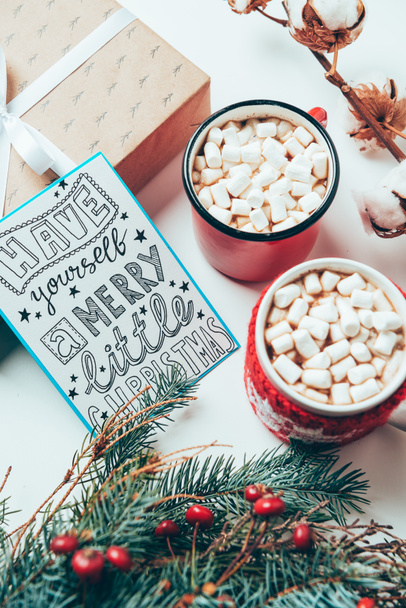 Draufsicht auf Postkarte mit selbst gemachtem Weihnachts-Schriftzug, Geschenk und Tassen mit Kakaogetränken mit Marshmallows auf weißer Tischplatte - Foto, Bild