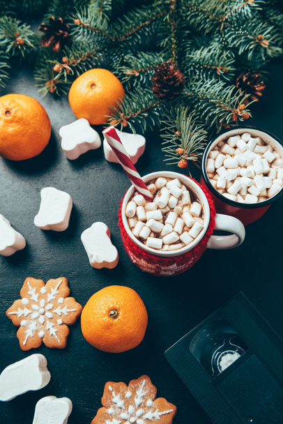 top view of cups of hot drinks with marshmallows, video cassette and tangerines on black surface, christmas breakfast concept - Fotografie, Obrázek
