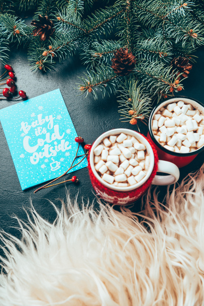 top view of cups of hot drinks with marshmallows, pine tree branches and postcard with baby its cold outside lettering on black surface, christmas and new year holidays concept - Photo, Image