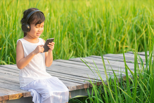 Asiática linda chica relajante sentado y el uso de teléfono móvil en el puente de madera en el campo de arroz en la puesta de sol con fondo de la naturaleza
. - Foto, Imagen