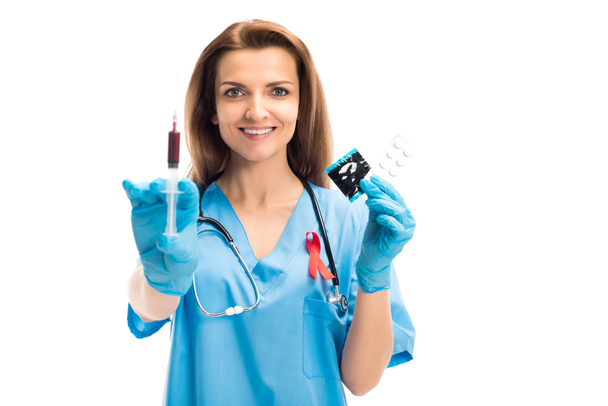 attractive smiling doctor with red ribbon holding syringe with blood and condom with pills isolated on white, world aids day concept - Foto, Imagen