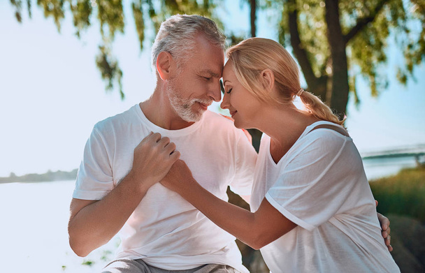 Senior couple is spending time together outdoors. Bearded handsome man and attractive woman are hugging while being in park. - Photo, image