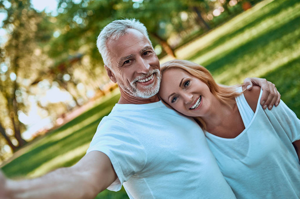 Senior couple is spending time together outdoors. Bearded handsome man and attractive woman are hugging while being in park. - Photo, Image