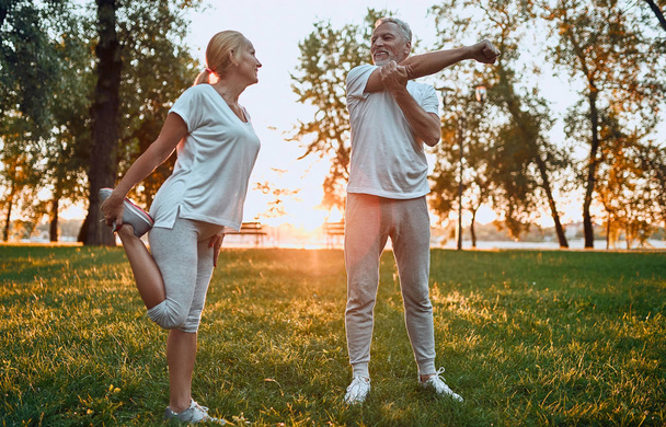 Senior couple is doing sport outdoors. Stretching in park during sunrise. - Фото, изображение