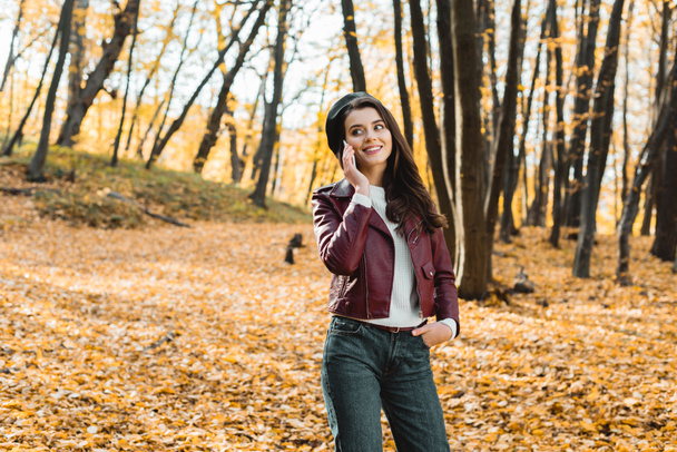 happy fashionable girl in leather jacket talking on smartphone in autumnal forest  - Fotografie, Obrázek
