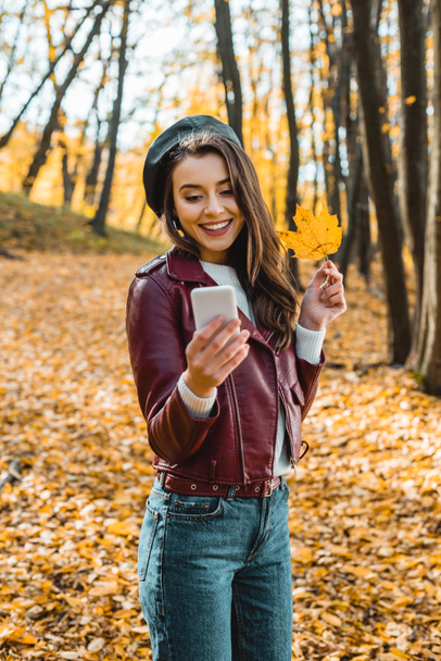 s úsměvem stylová holka v kožené bundě s selfie s žlutý list na smartphone venku - Fotografie, Obrázek